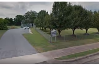 A paved driveway leading to a building partially visible in the background, with a grassy area and several trees on the right side. There is a sign near the trees and a sidewalk along the road in the foreground.