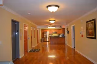 A long hallway with warm wood floor leads to a kitchen in a senior living facility.