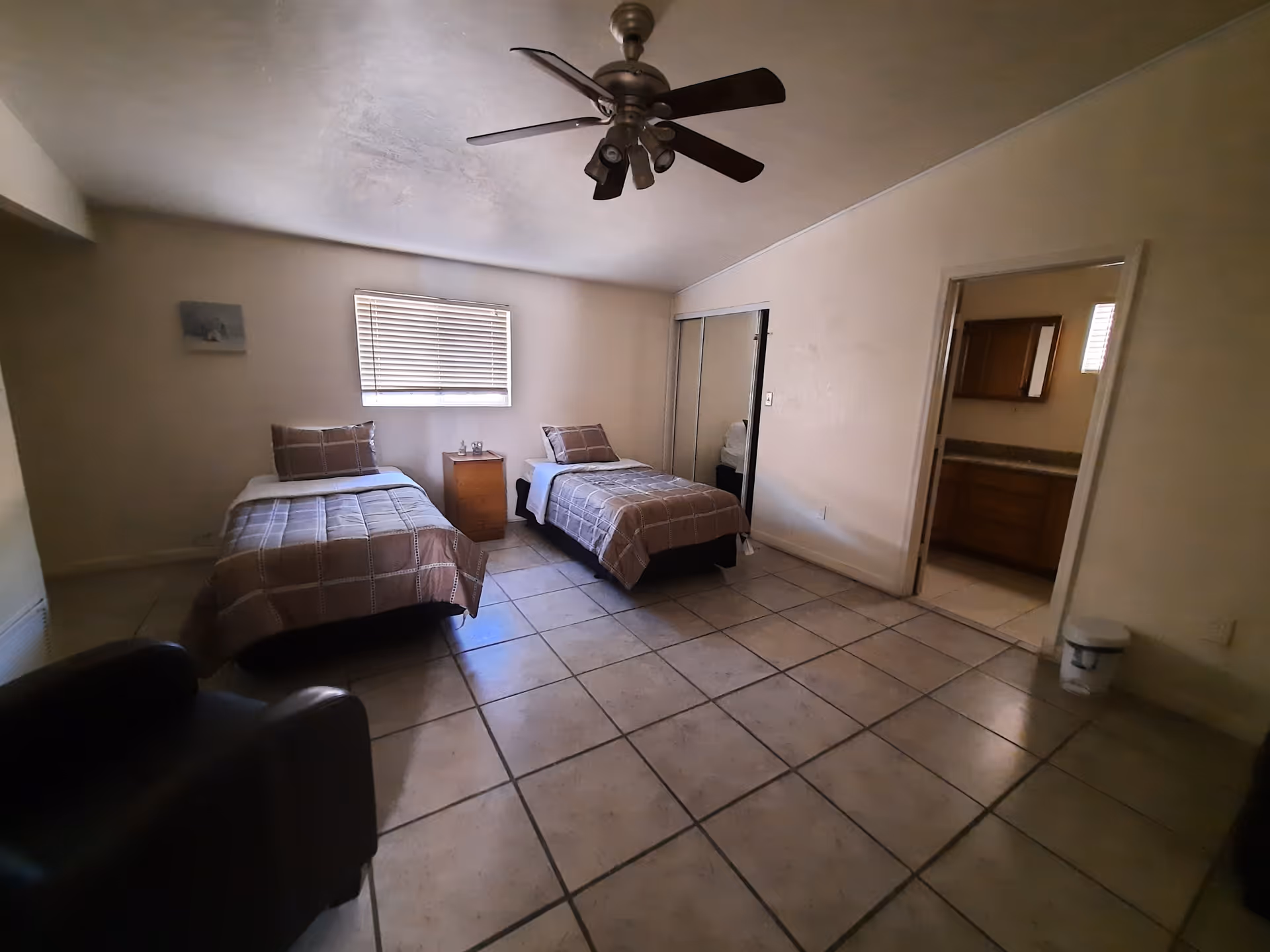 A bedroom with two single beds covered with brown checkered bedspreads, a small wooden nightstand between the beds, a window with blinds above the nightstand, a ceiling fan with lights, a mirrored closet door, a doorway leading to a bathroom with a wooden cabinet and granite countertop, and a dark armchair in the corner.
