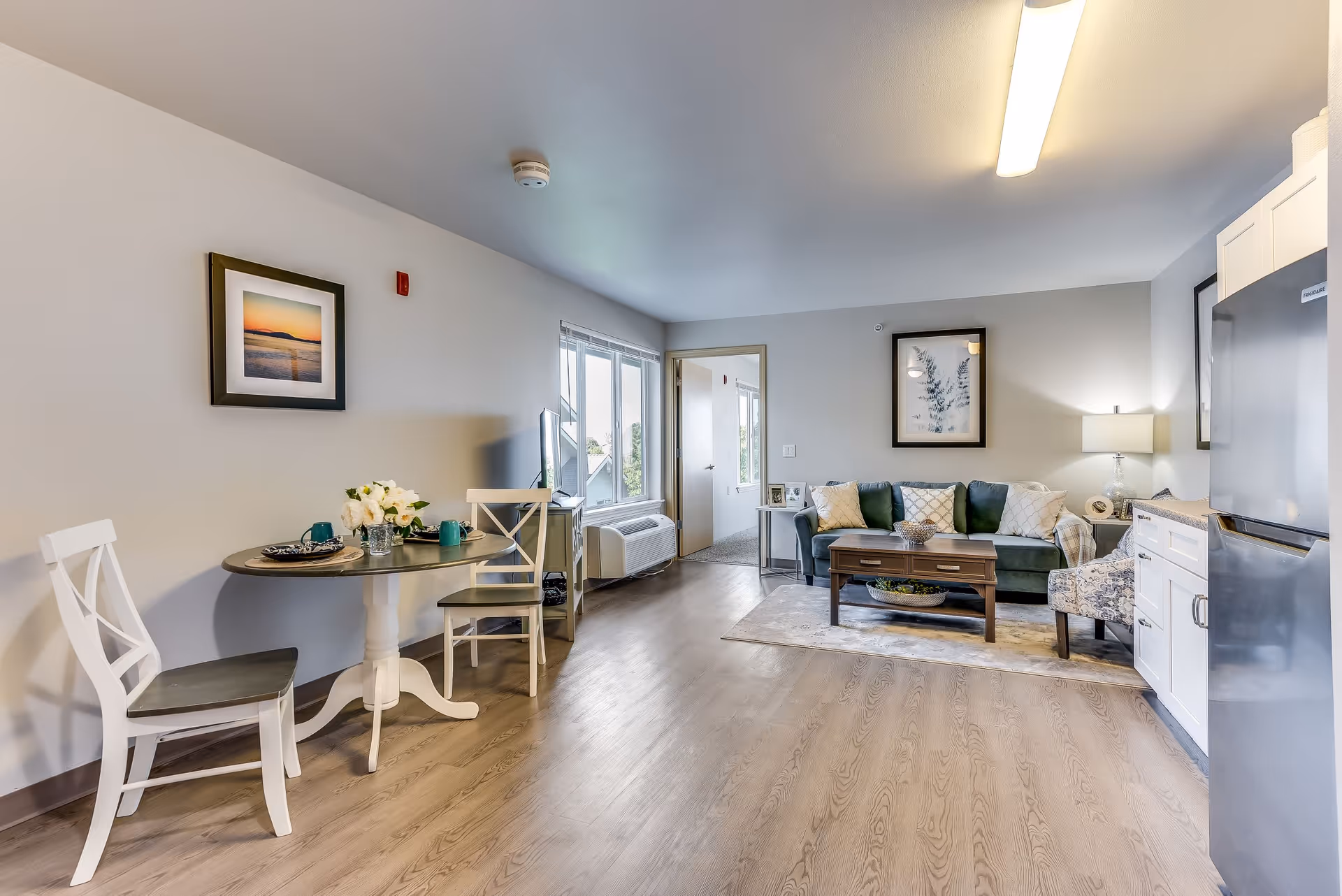 A bright and clean senior living apartment interior featuring a small round dining table with two white chairs on the left, a green couch with decorative pillows, a wooden coffee table, and a patterned armchair on the right. The room has light wood flooring, a large window, framed artwork on the walls, and a kitchen area with white cabinets and a stainless steel refrigerator on the far right.