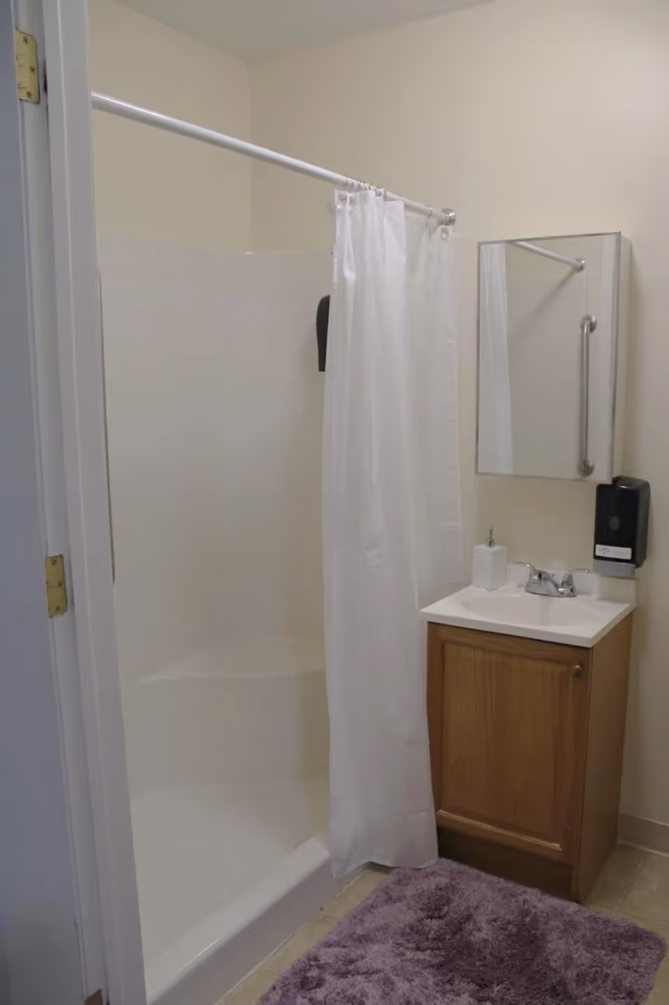 A small bathroom with a white shower stall and a white shower curtain on a curved rod. Next to the shower is a wooden vanity with a white sink and a silver faucet. Above the sink is a rectangular mirror with a grab bar mounted on the wall beside it. A soap dispenser is mounted on the wall next to the sink. A purple bath mat is on the floor in front of the vanity.