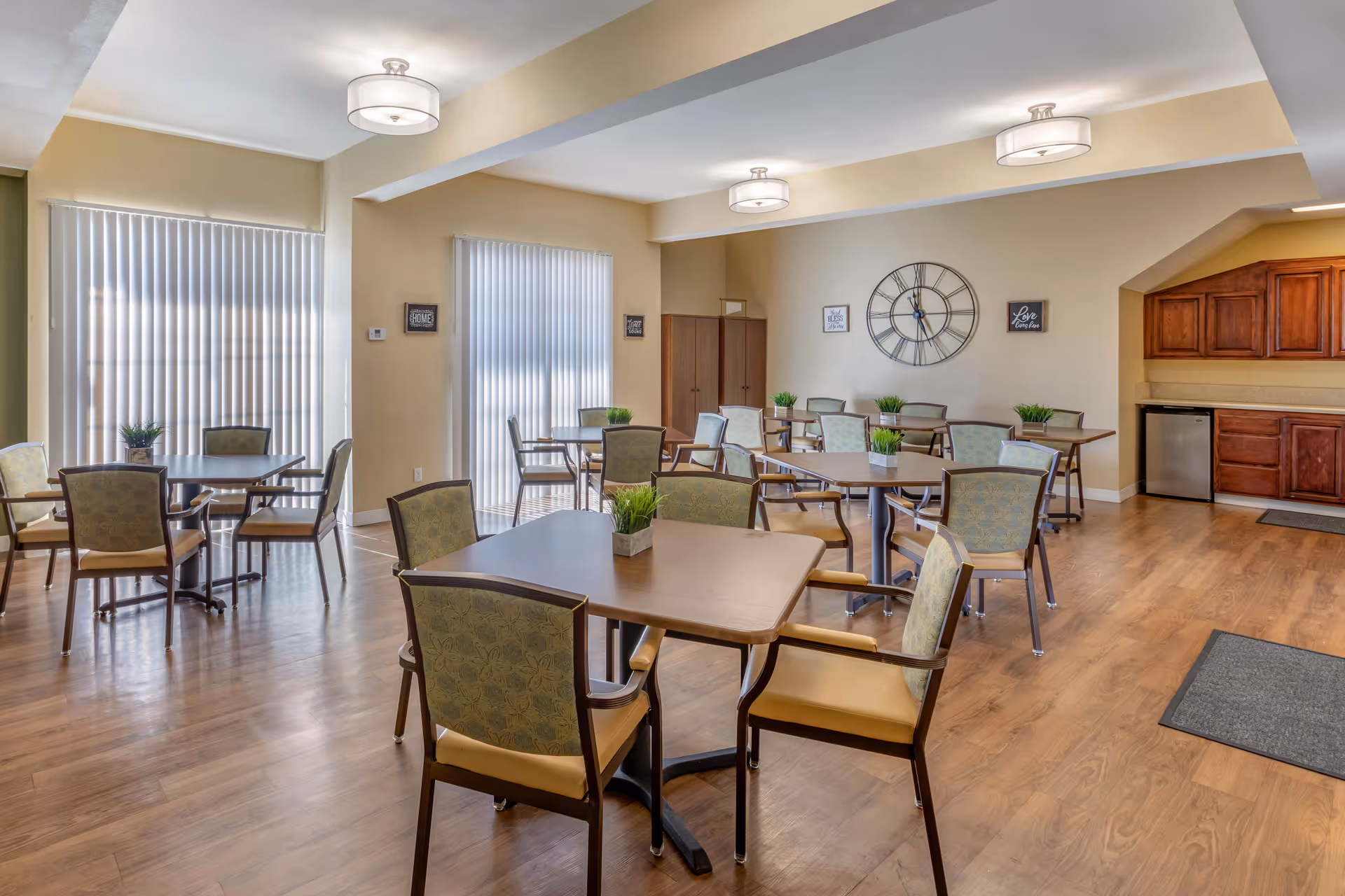 A bright dining room with several square wooden tables, each surrounded by four cushioned chairs with patterned backs. Small green plants in rectangular pots are placed on each table. The room has light beige walls, wood flooring, and three ceiling lights. Vertical blinds cover the windows, and a large decorative clock and framed signs hang on the walls. There is a small kitchenette area with wooden cabinets and a mini refrigerator in the corner.