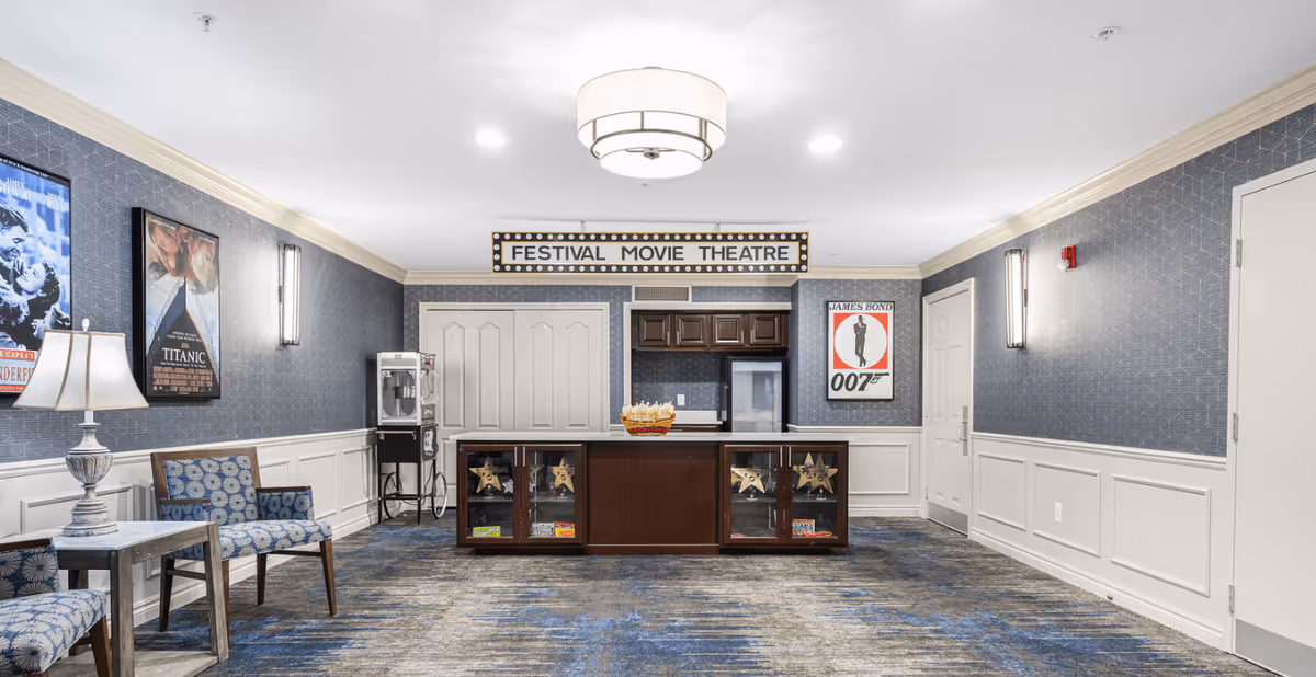 Interior view of a movie theatre lounge area at Waltonwood Twelve Oaks featuring a popcorn machine, a counter with glass display cabinets, movie posters on the walls including Titanic and James Bond 007, blue patterned carpet, and seating with blue patterned chairs and a small table with a lamp.