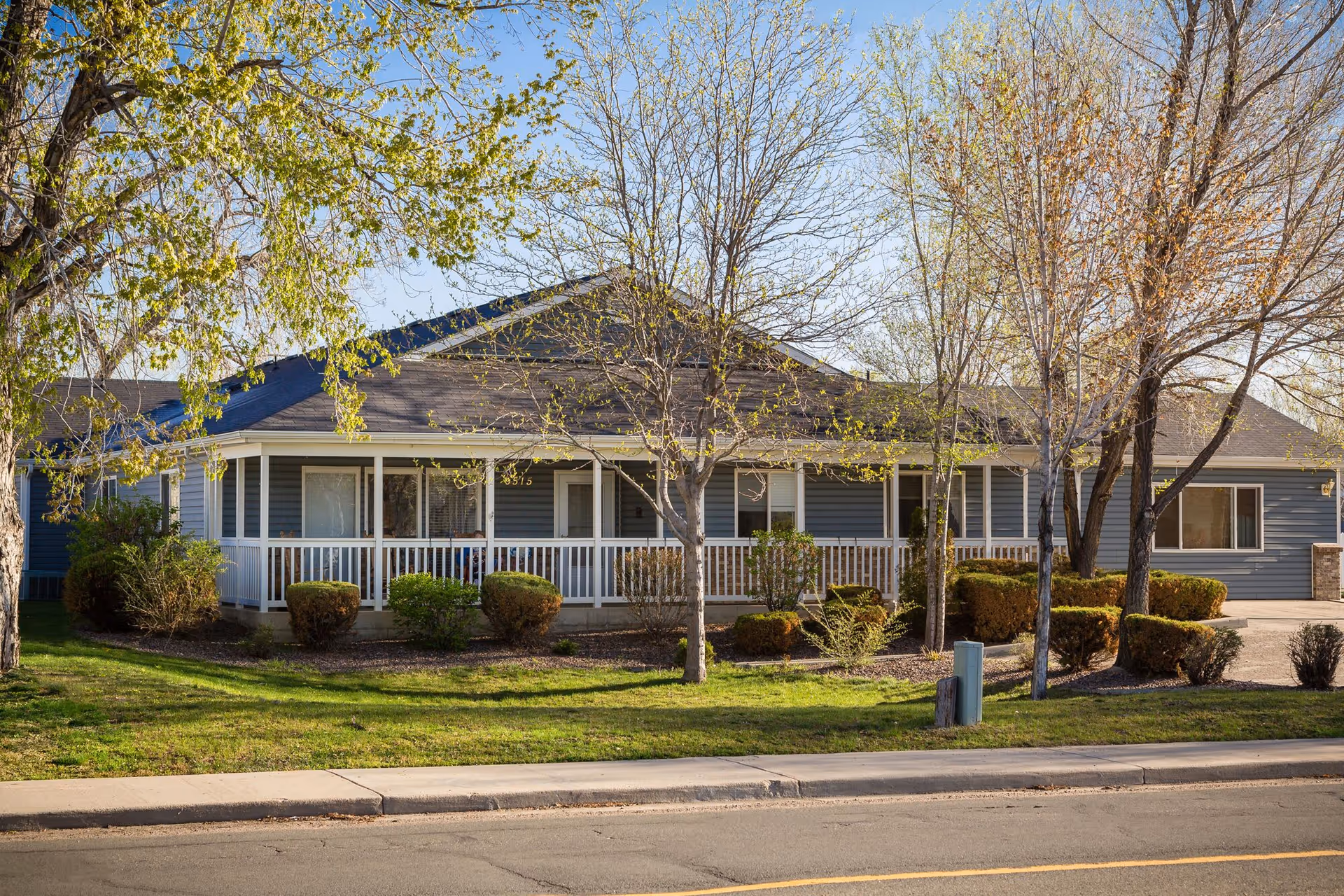 Exterior view of a single-story assisted living facility building with a covered porch, surrounded by trees and shrubs, under a clear blue sky.