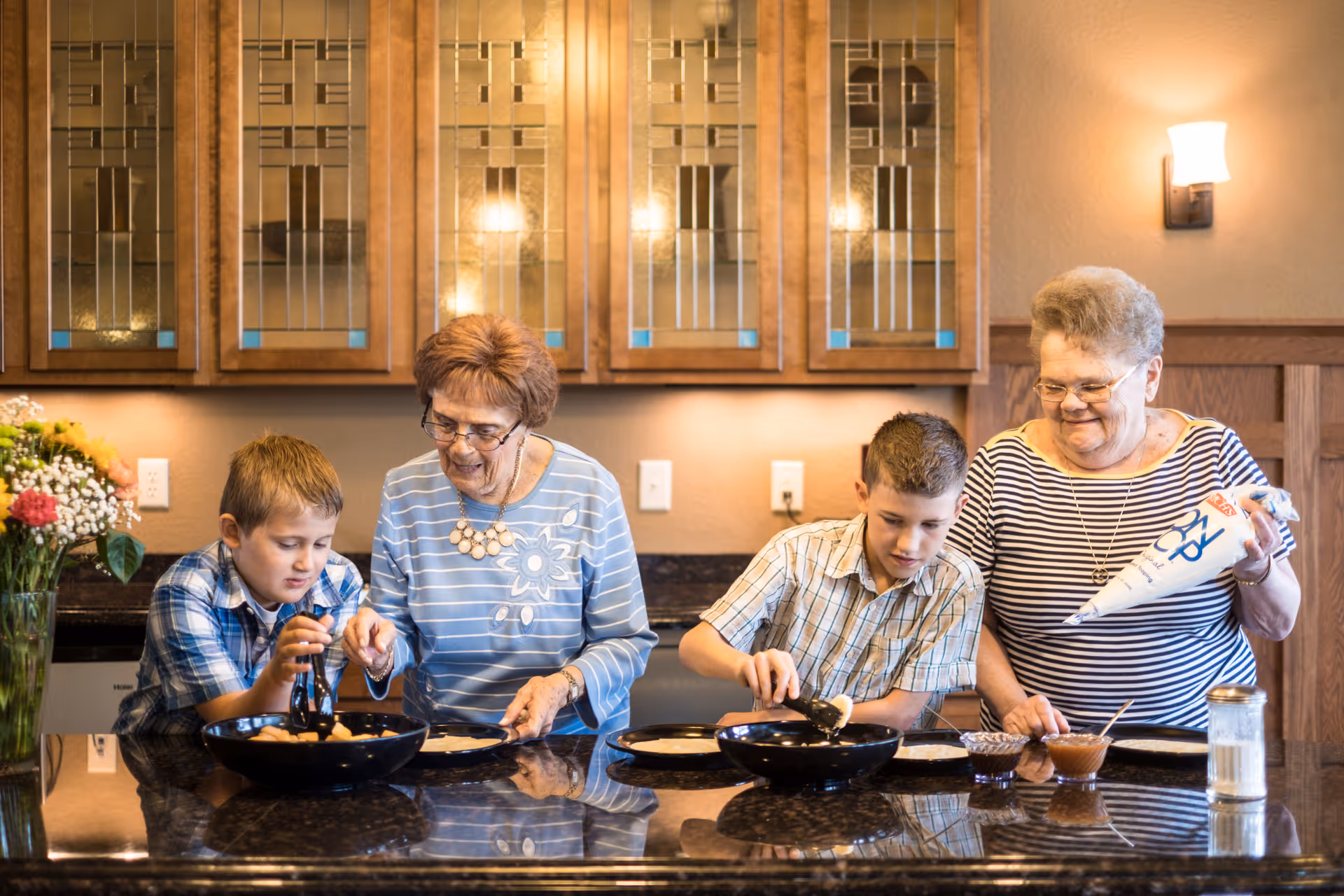 Two elderly women and two young boys preparing food together at a kitchen counter.