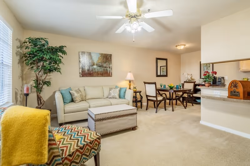 A bright and cozy living room and dining area in an independent living facility. The living room features a cream-colored sofa with decorative pillows, a patterned armchair with a yellow throw blanket, a large potted plant, and a ceiling fan with lights. The dining area has a wooden table set with four chairs, blue glassware, and a bowl of fruit. The room has beige walls and carpet, with a painting and a mirror on the walls, and a small kitchen counter visible in the background.