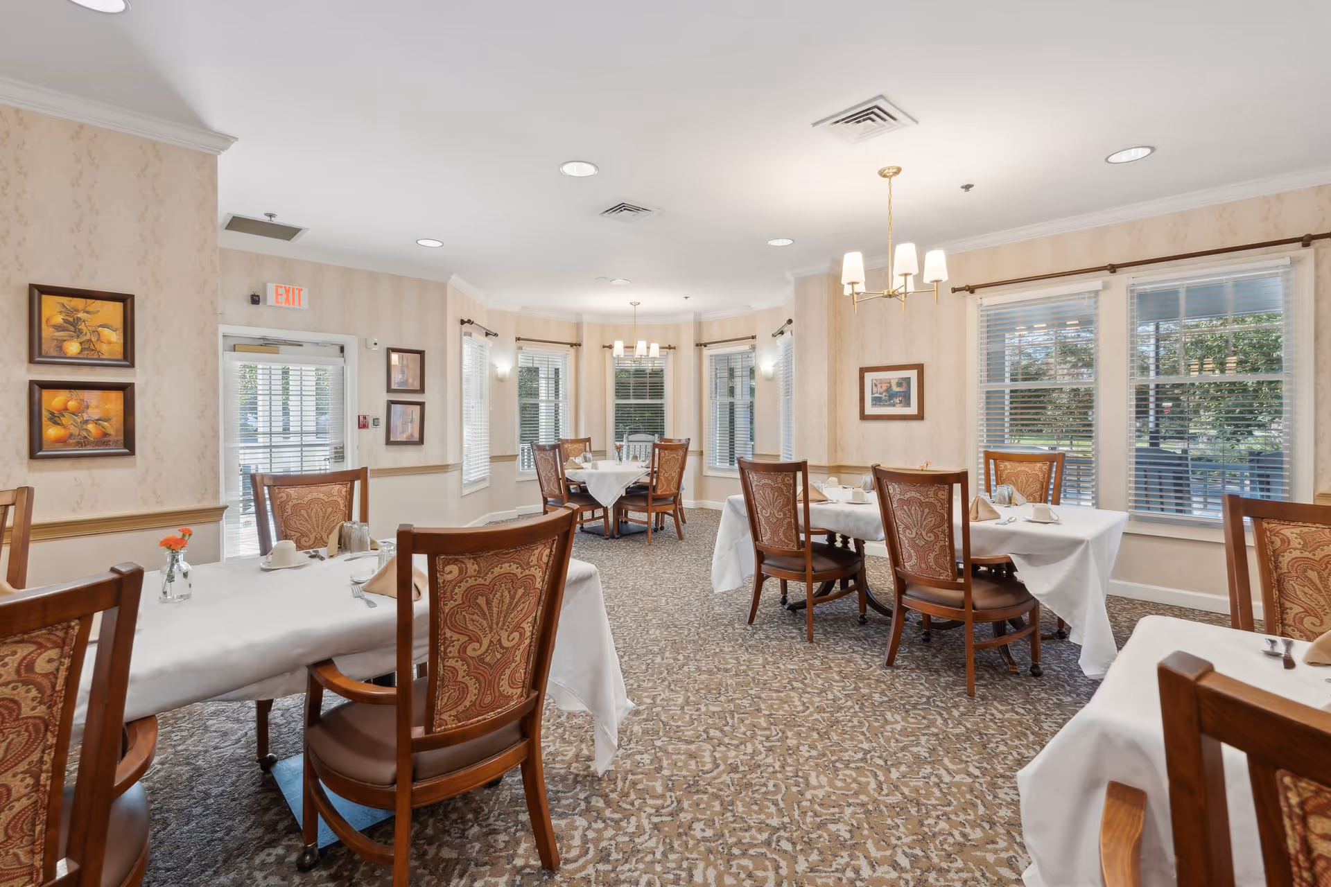 A bright dining room with several tables covered with white tablecloths, each set with cups, napkins, and silverware. The room features patterned carpet, beige wallpaper, wooden chairs with ornate upholstery, and multiple windows with white blinds letting in natural light. There are framed pictures on the walls and ceiling light fixtures providing additional illumination.