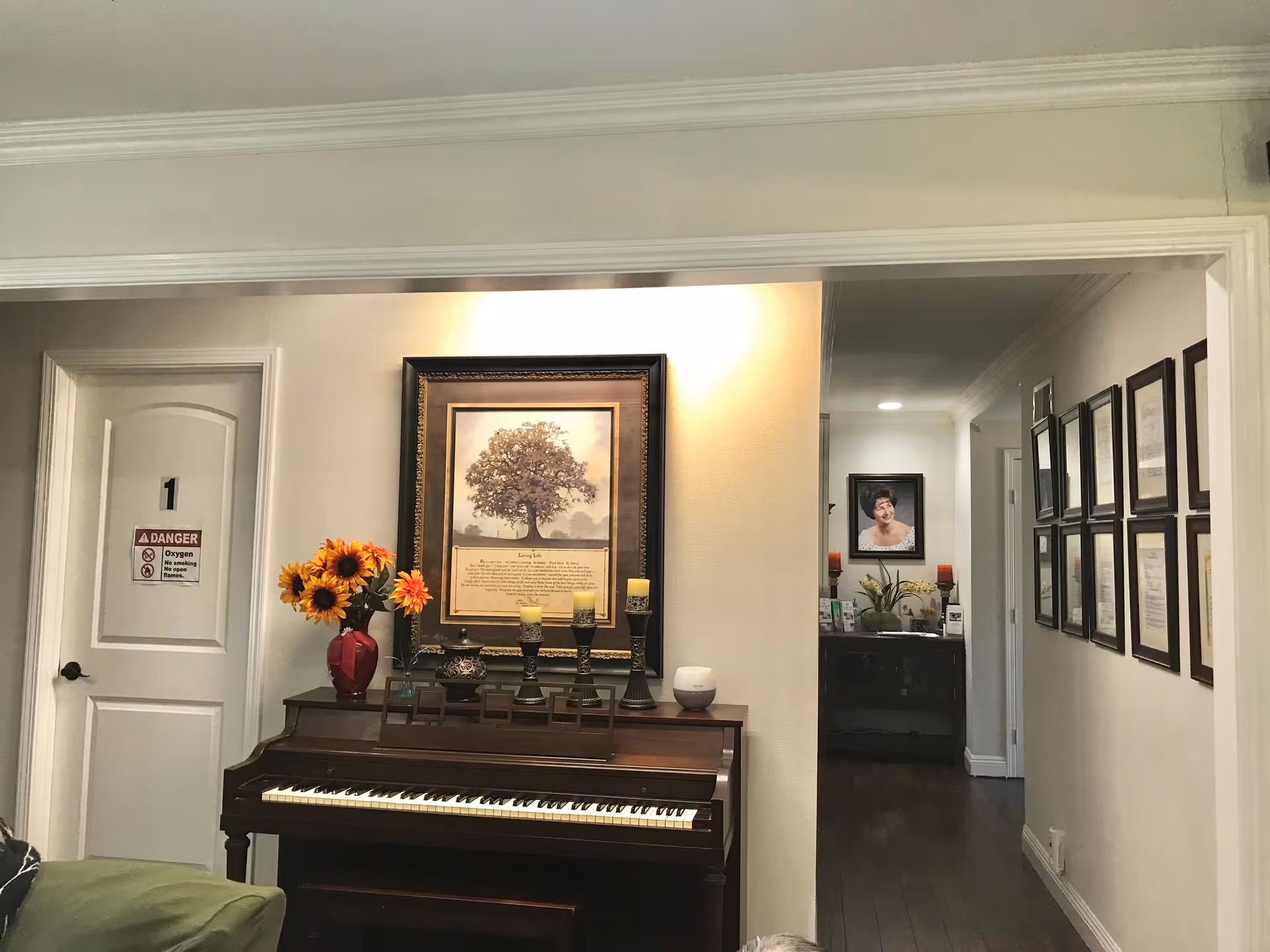 Interior view of a room in Mcfadden Assisted Living showing a wooden piano with decorative candles and a vase of sunflowers on top. Above the piano is a framed picture of a tree with a poem. To the left is a white door labeled with a danger sign warning about oxygen and no open flames. In the background, a hallway with framed certificates on the right wall and a portrait of a woman at the end of the hall is visible.