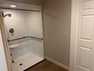 A clean, accessible shower area with a white shower stall featuring a built-in seat and grab bars. The shower has a handheld showerhead and a control knob. The surrounding walls are painted beige, and there is a white door to the right side of the image.