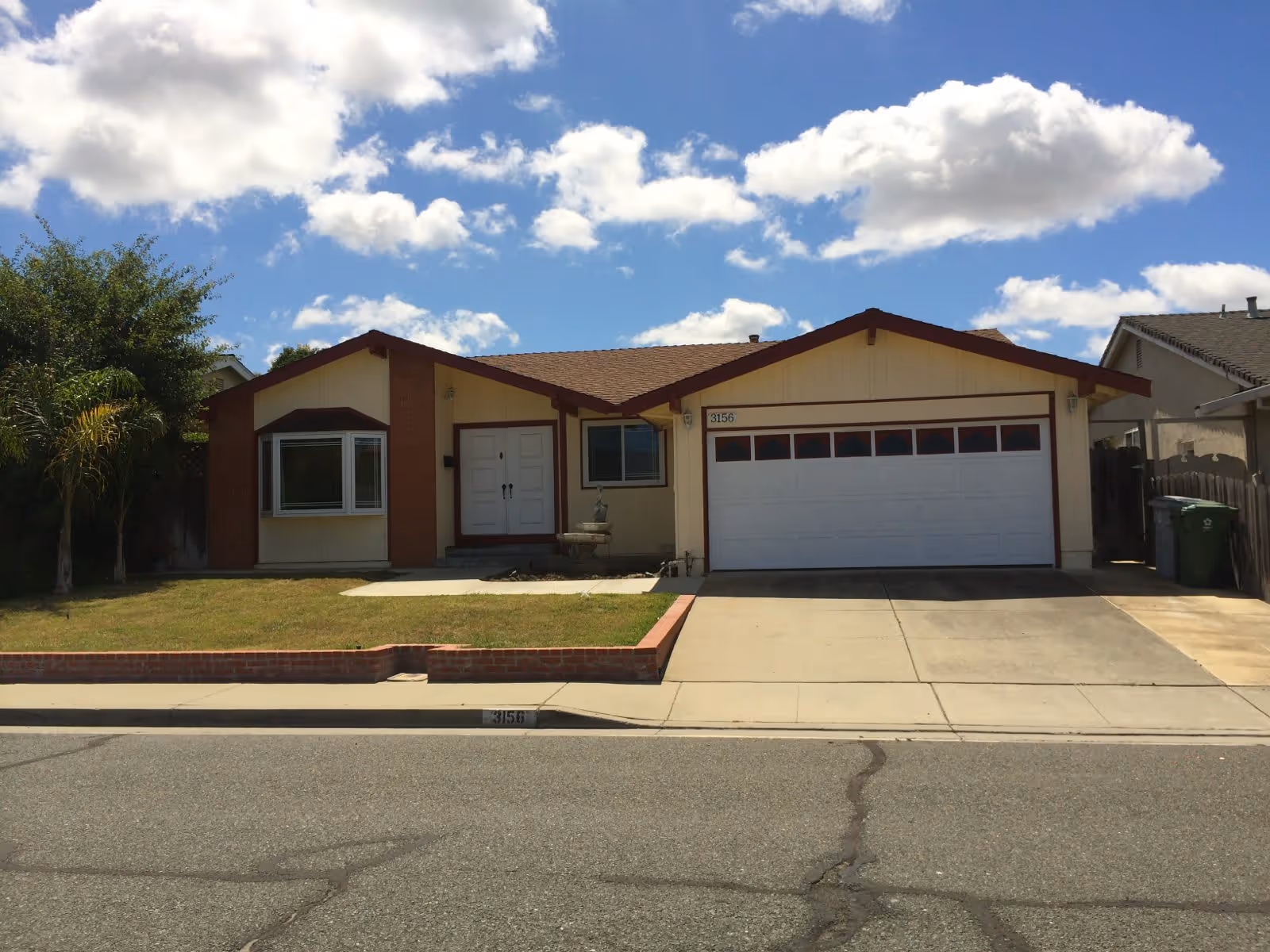 Single-story house with a beige exterior and brown trim, featuring a double garage door, a front door with two panels, a bay window on the left, a small lawn with a brick border, and a driveway. The sky is blue with scattered white clouds.