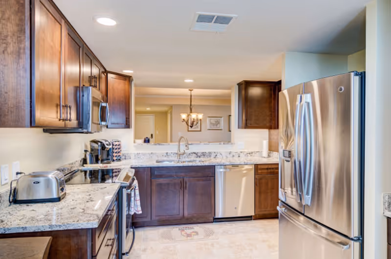 Modern kitchen with dark wooden cabinets, granite countertops, stainless steel refrigerator, dishwasher, stove, and toaster. The kitchen has recessed lighting and a view into a dining area with a chandelier.
