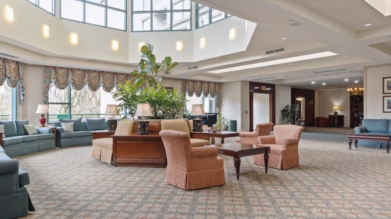 Spacious and well-lit living room area with large windows, multiple seating arrangements including sofas and armchairs, a central wooden table with lamps and plants, and a high ceiling with skylights.