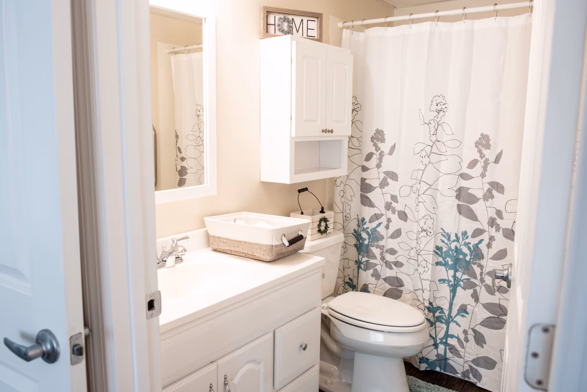 Bright small bathroom with a white vanity and mirror, a toilet, wall cabinet, and a floral shower curtain.