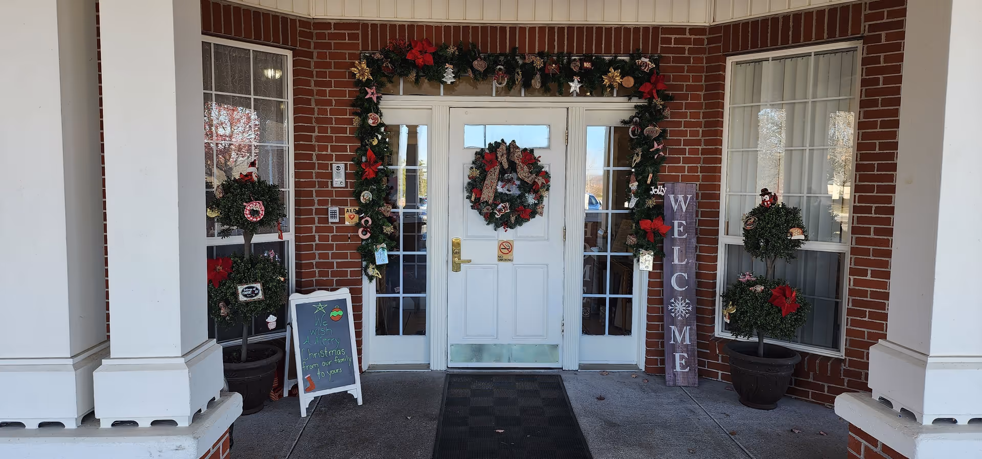 Entrance of Wellington Manor Assisted Living decorated for Christmas with a wreath on the white door, garland around the door frame, potted plants with holiday decorations on either side, and a wooden welcome sign. There is also a small chalkboard sign with holiday messages near the left side.