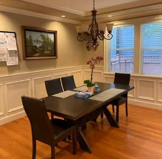 A dining room with a dark wooden table and four black chairs. The table has placemats, a tissue box, and a potted orchid. The room has wooden flooring, beige walls with white wainscoting, a chandelier hanging above the table, a framed landscape painting on the wall, and two windows with blinds partially open.