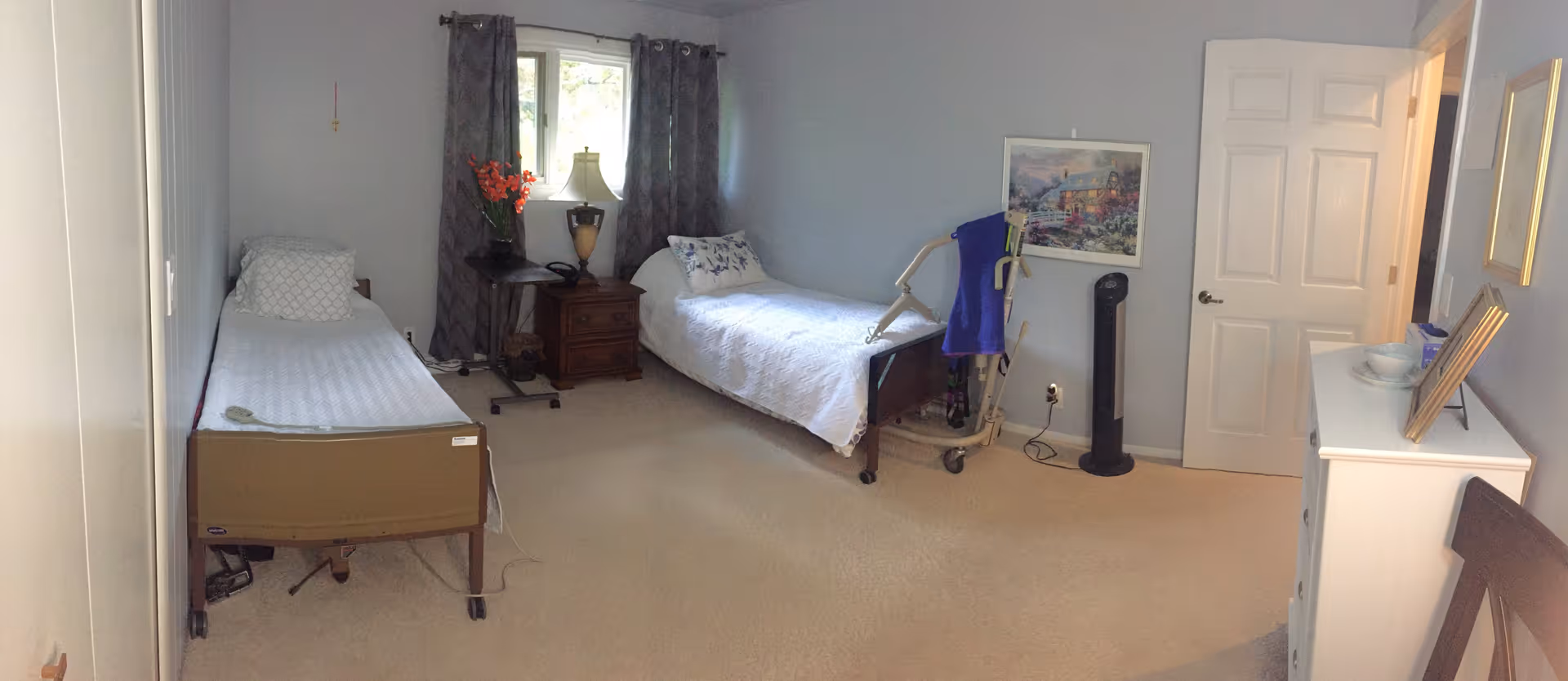 A bedroom with two single beds, each with white bedding and pillows. Between the beds is a wooden nightstand with a lamp, a phone, and a vase with orange flowers. A window with gray curtains is behind the nightstand. On the right side of the room, there is a white dresser with a bowl and picture frames on top, a standing fan, and a walker with a blue cloth draped over it. A framed painting hangs on the wall above the walker. The room has beige carpet and light-colored walls.
