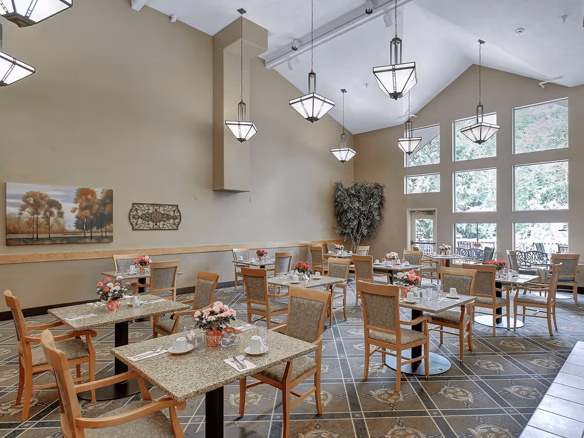 A spacious dining room with multiple tables and chairs arranged neatly. Each table has a small floral centerpiece and place settings with cups, saucers, and utensils. Large windows on one side let in natural light, and modern pendant lights hang from the high ceiling. The walls are painted beige and decorated with a landscape painting and a decorative metal piece. There is a potted plant in the corner near the windows.
