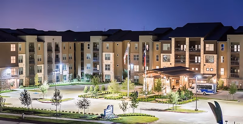 Illuminated multi-story senior living building at night with a landscaped circular driveway, flagpoles, and a covered main entrance.
