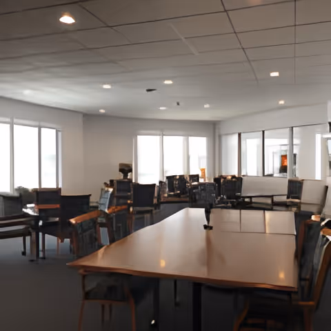 A spacious dining room with multiple wooden tables and chairs arranged neatly. Large windows allow natural light to fill the room, and the ceiling has recessed lighting. The room has a clean and organized appearance.