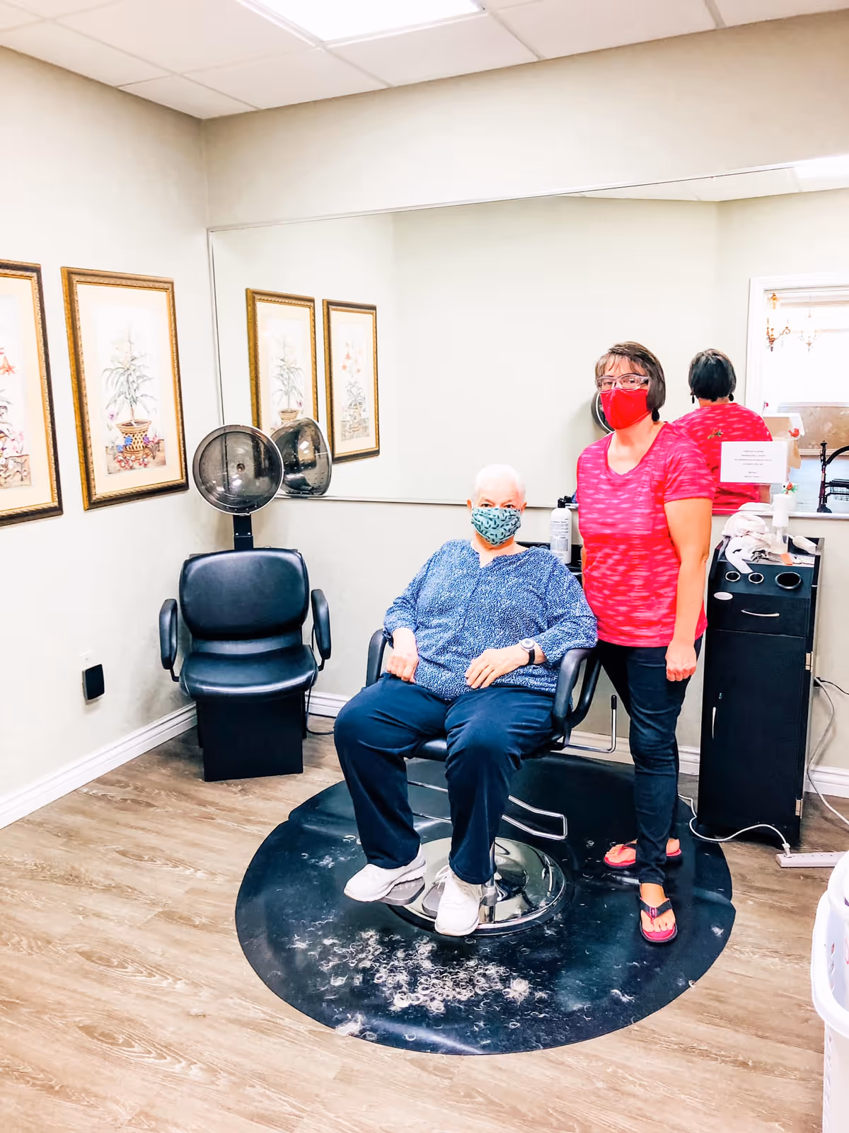 A senior woman wearing a blue patterned top and a face mask is seated in a salon chair inside a hair salon area. Next to her stands a woman in a red patterned shirt and face mask. The room has light-colored walls with three framed botanical prints, a large mirror, a black salon chair with a hair dryer, and wood flooring.