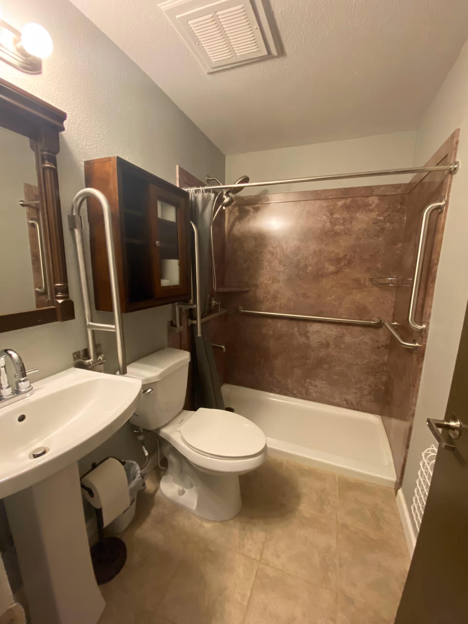 A small bathroom with a white pedestal sink, a toilet, and a bathtub with a shower curtain. The bathtub area has brown textured wall panels and multiple grab bars for accessibility. There is a wooden cabinet mounted on the wall above the toilet and a mirror above the sink. The floor is tiled in a light beige color.