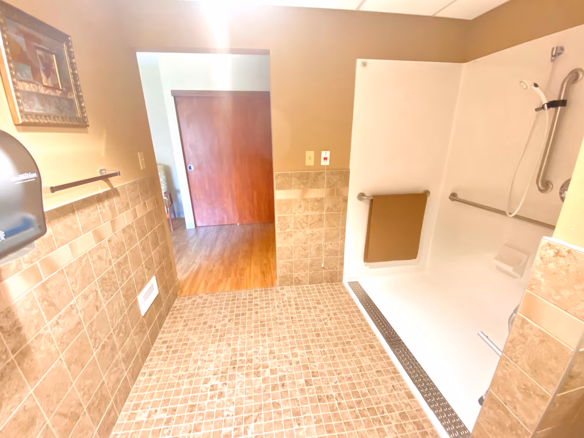 Accessible bathroom with tiled floor, roll-in shower with grab bars and a fold-down seat, and a doorway leading to a room with wood flooring.