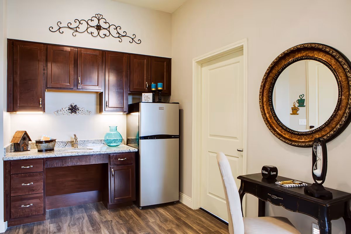 A small kitchenette area with dark wooden cabinets, a granite countertop, a stainless steel mini refrigerator, and a sink. Decorative items include a birdhouse, a glass vase, and candles. To the right, there is a black wooden desk with a round ornate mirror above it, a white upholstered chair, and small decorative objects on the desk. The floor is wood-style laminate, and the walls are painted light beige.
