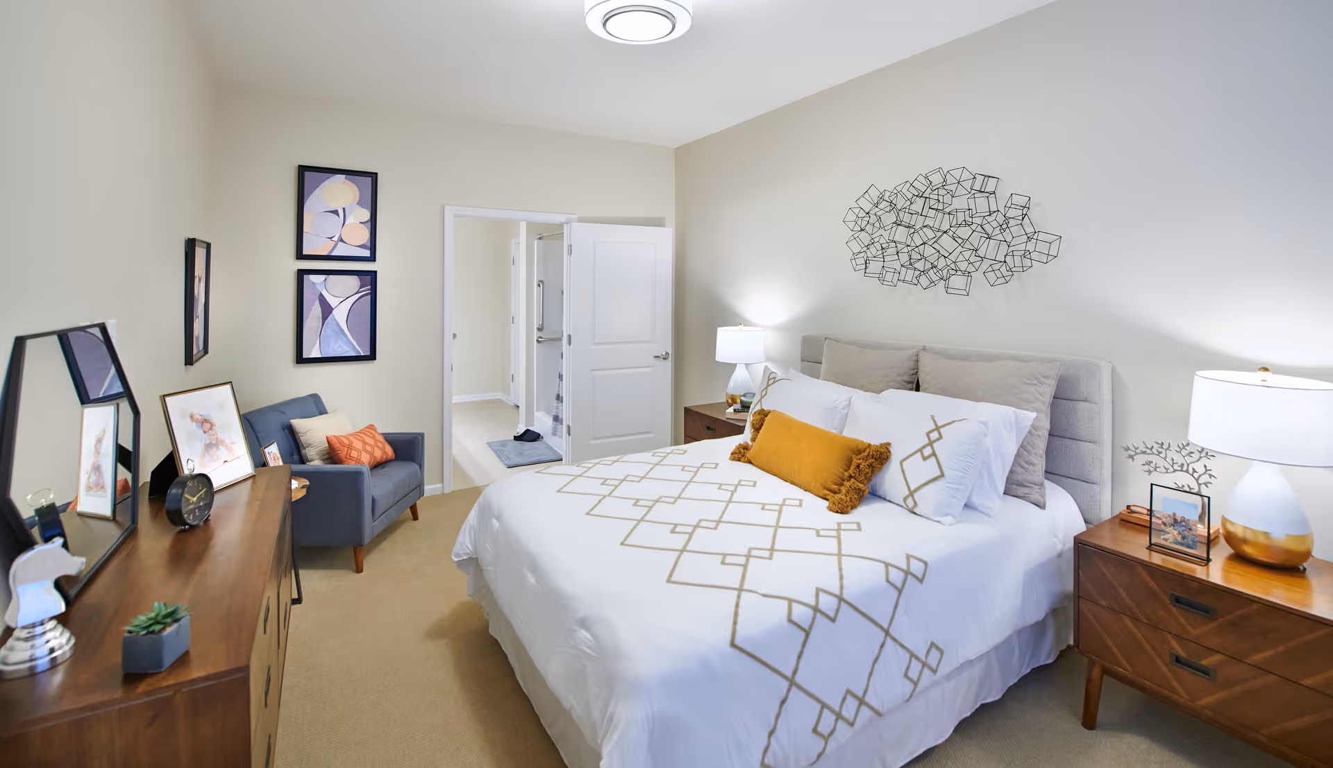 A neatly arranged bedroom with a large bed featuring white bedding with a geometric pattern and a mustard yellow accent pillow. There are two wooden nightstands with lamps on either side of the bed. A decorative metal wall art piece hangs above the bed. To the left, there is a wooden dresser with a mirror, framed pictures, and a small plant. A gray armchair with cushions is positioned against the wall, which has two framed abstract art pieces. The door to an adjoining bathroom is open, showing a glimpse of the shower area.