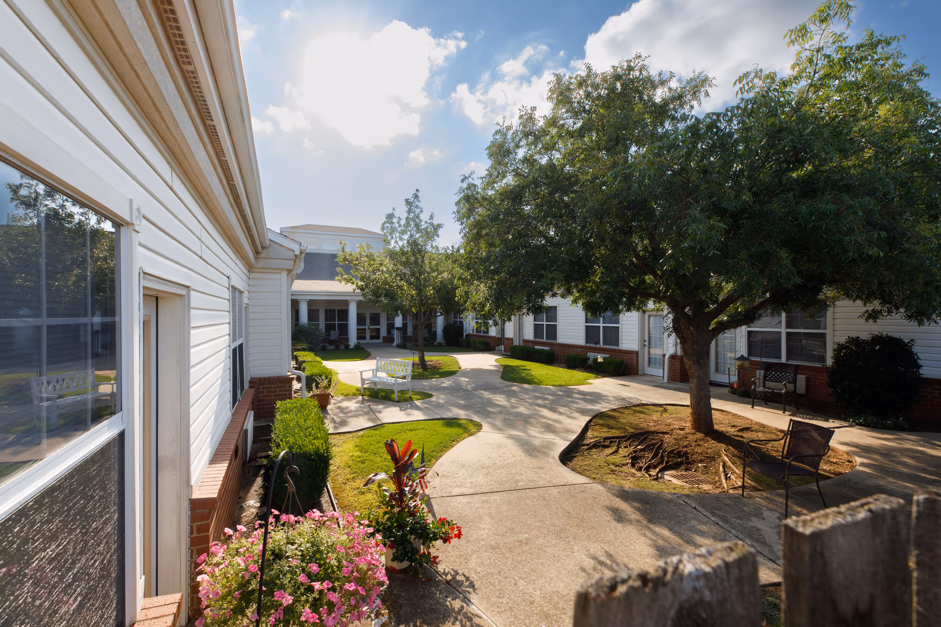 A sunny courtyard area at Arbor House of Midwest City featuring a concrete walkway winding through green grass, trees, and flower beds. White benches and chairs are placed along the path near the building entrances with white siding and brick accents. The sky is partly cloudy with sunlight casting shadows on the ground.