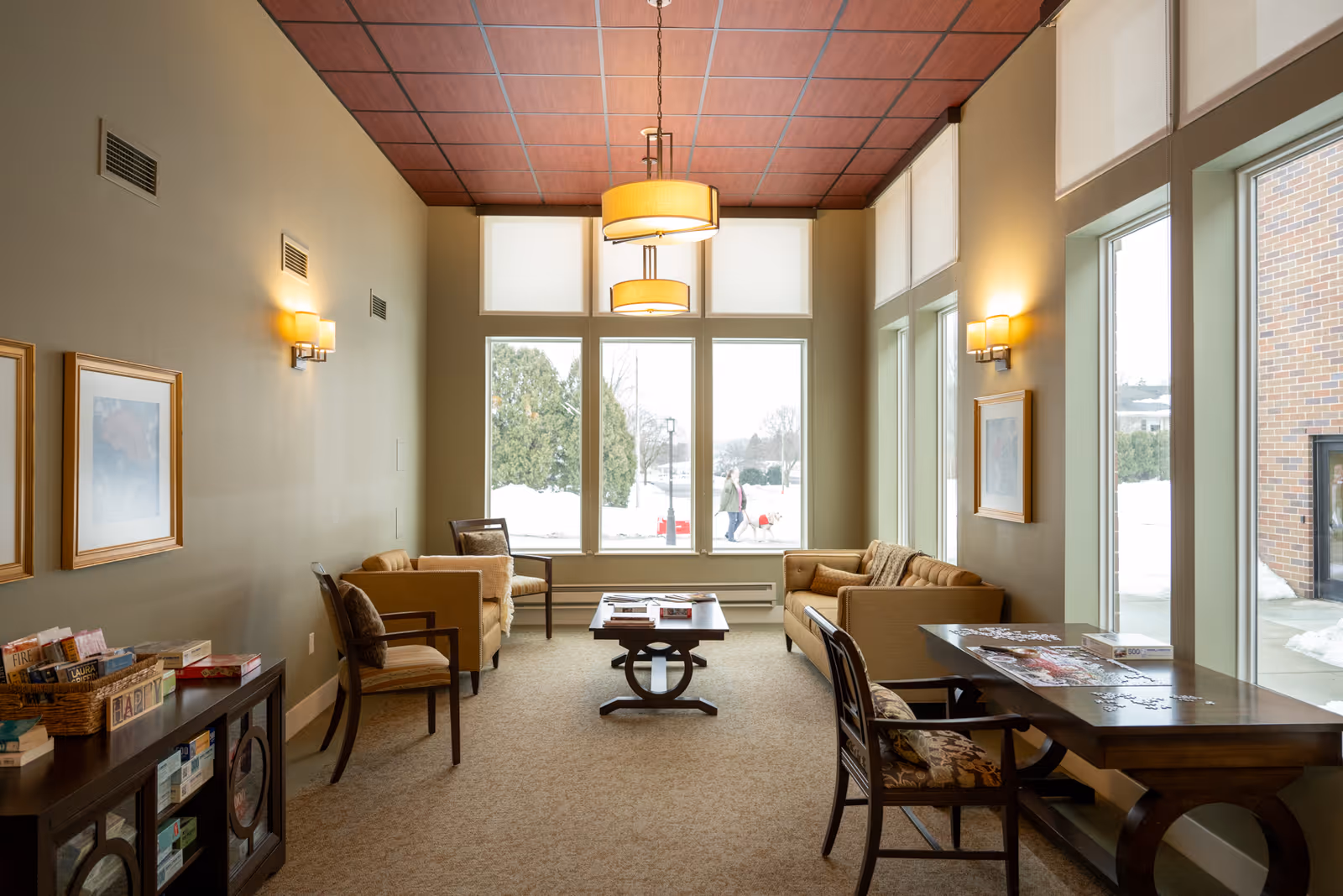 A bright senior living lounge with sofas, chairs, a coffee table and large windows overlooking a snowy exterior.