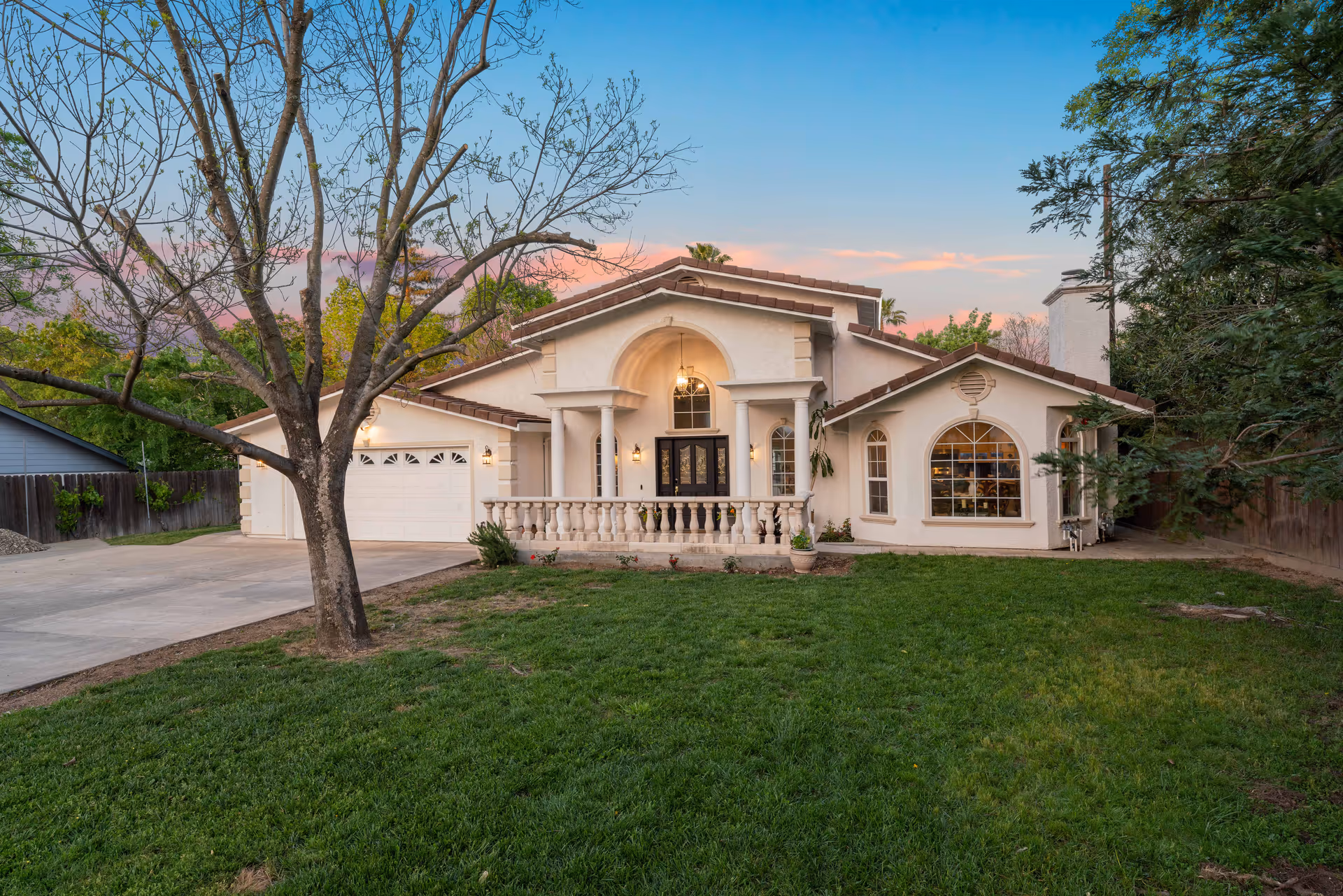 Front exterior view of a single-story house with a large green lawn, a leafless tree in the foreground, a driveway leading to a two-car garage, and a covered porch with columns and a black front door under a clear sky at sunset.