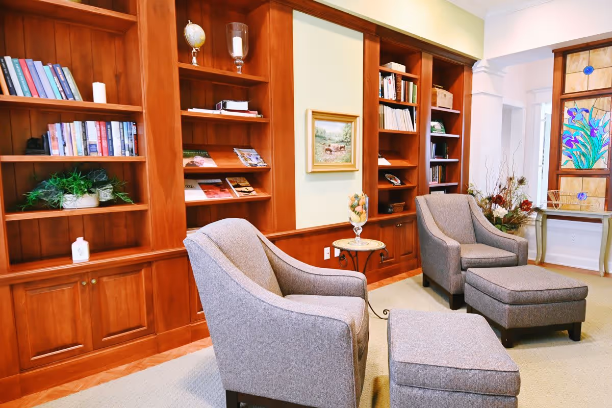 A cozy sitting area with two gray upholstered armchairs and matching ottomans in front of wooden built-in bookshelves filled with books, plants, and decorative items. A small round side table with a glass vase holding decorative fruit is placed between the chairs. The room features a light green wall with a framed painting and a stained glass window with floral designs.