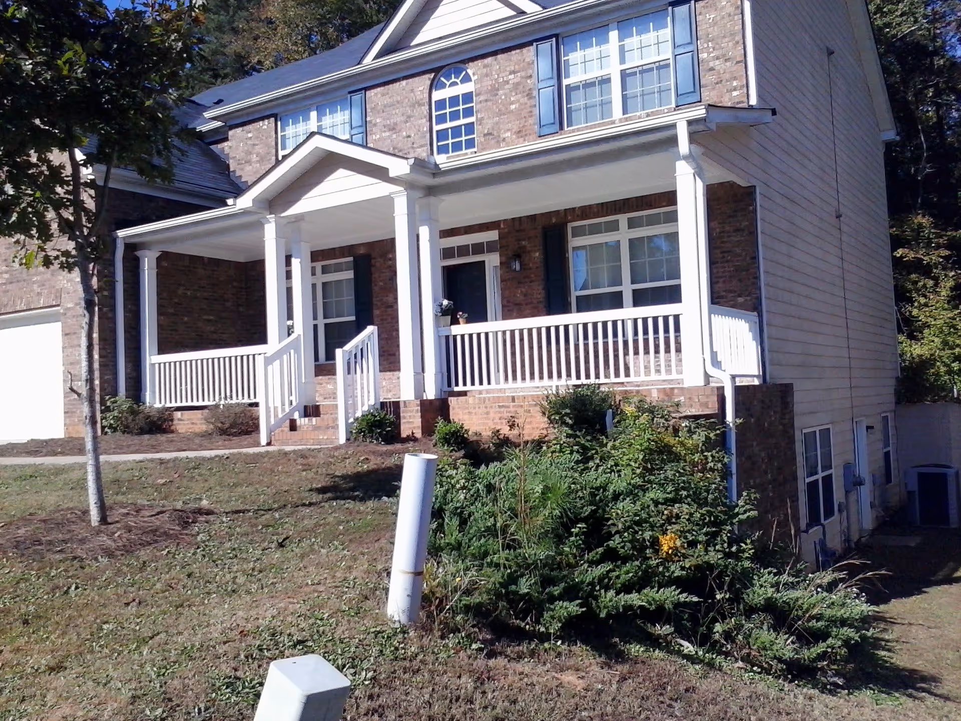 Front exterior view of a two-story brick house with white columns and railings on the porch, multiple windows with blue shutters, a small tree and bushes in the front yard, and a garage on the left side.