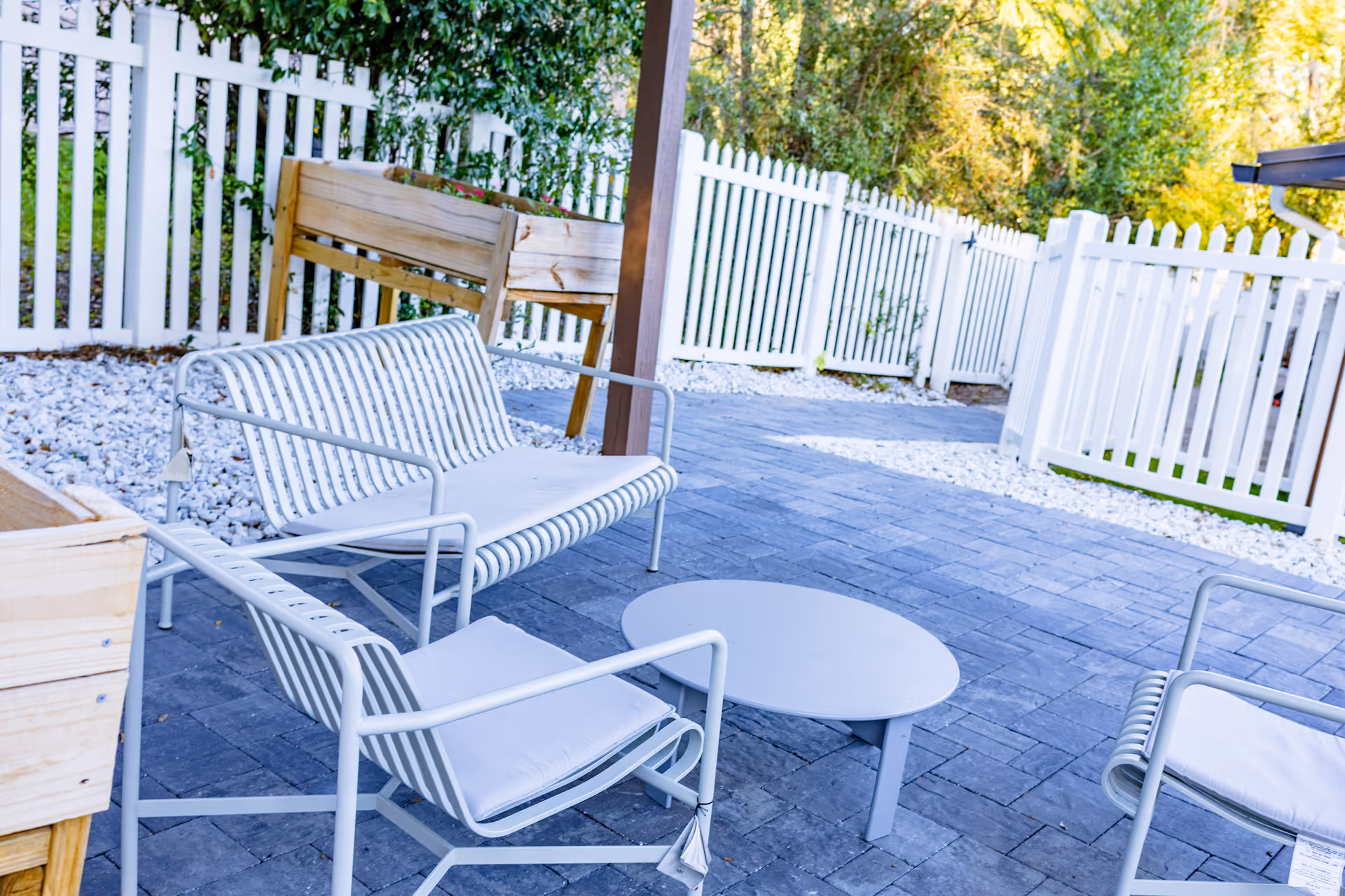 Outdoor patio area with white metal chairs and a matching loveseat around a low round table, surrounded by a white picket fence and greenery in the background.