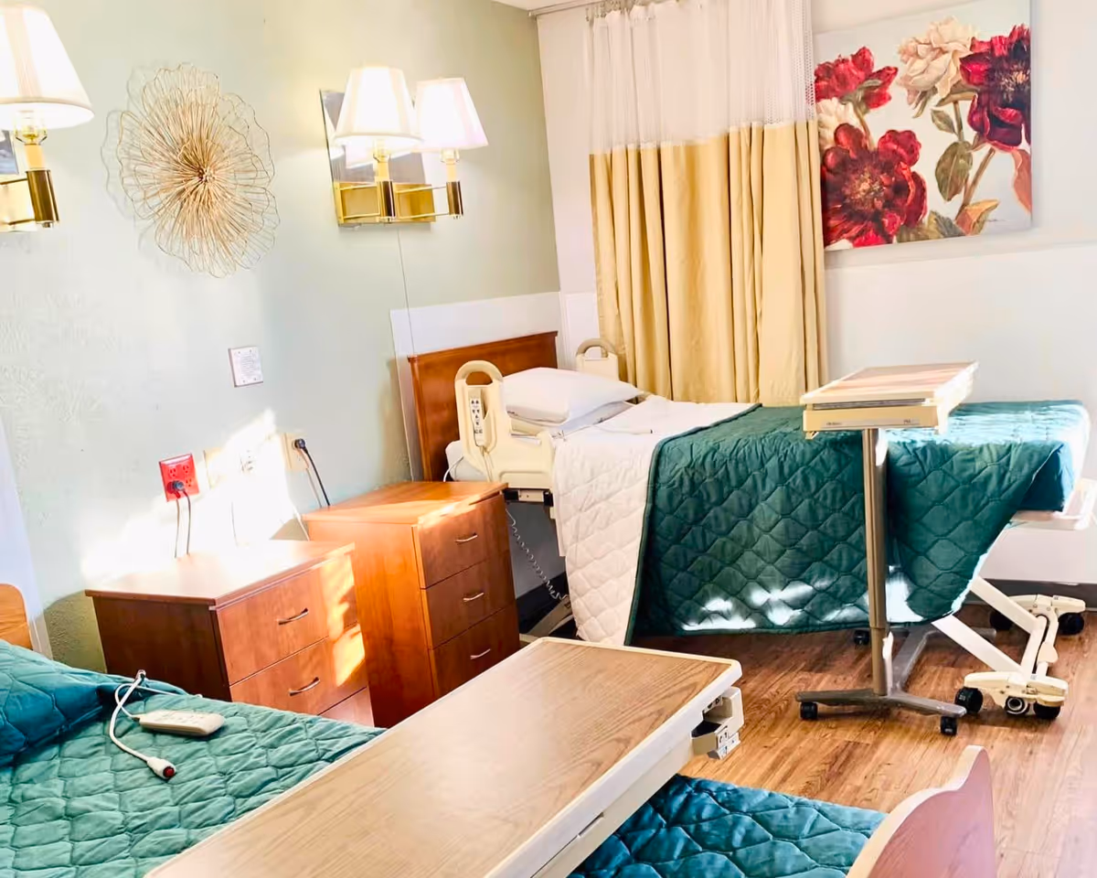 A bright senior care bedroom with two hospital-style beds, wooden nightstands, and green quilted bedcovers.