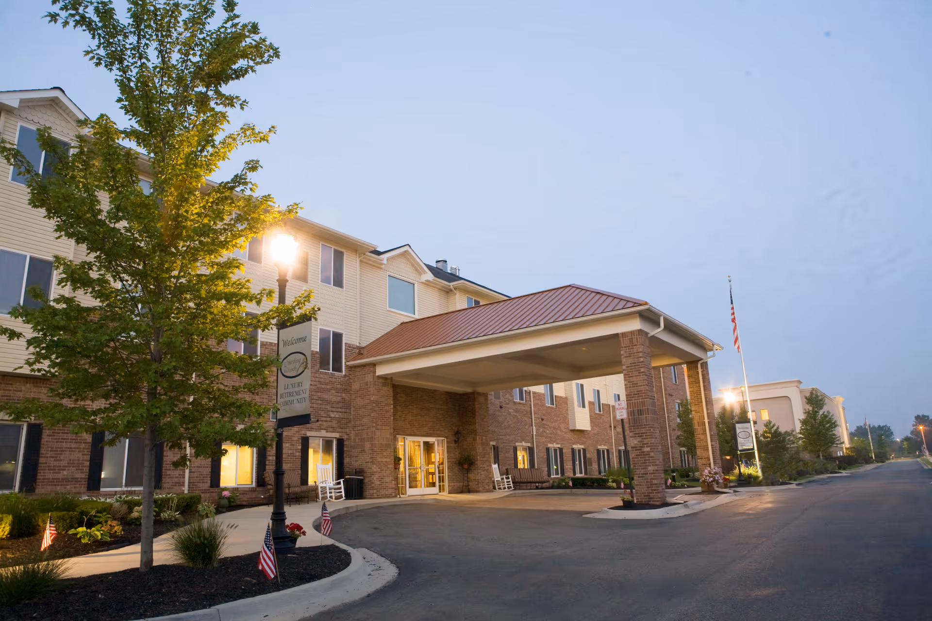 Exterior view of the American House Sterling Woods senior living facility at dusk, showing a three-story building with a covered entrance, a tree with a streetlamp, and small American flags along the sidewalk.