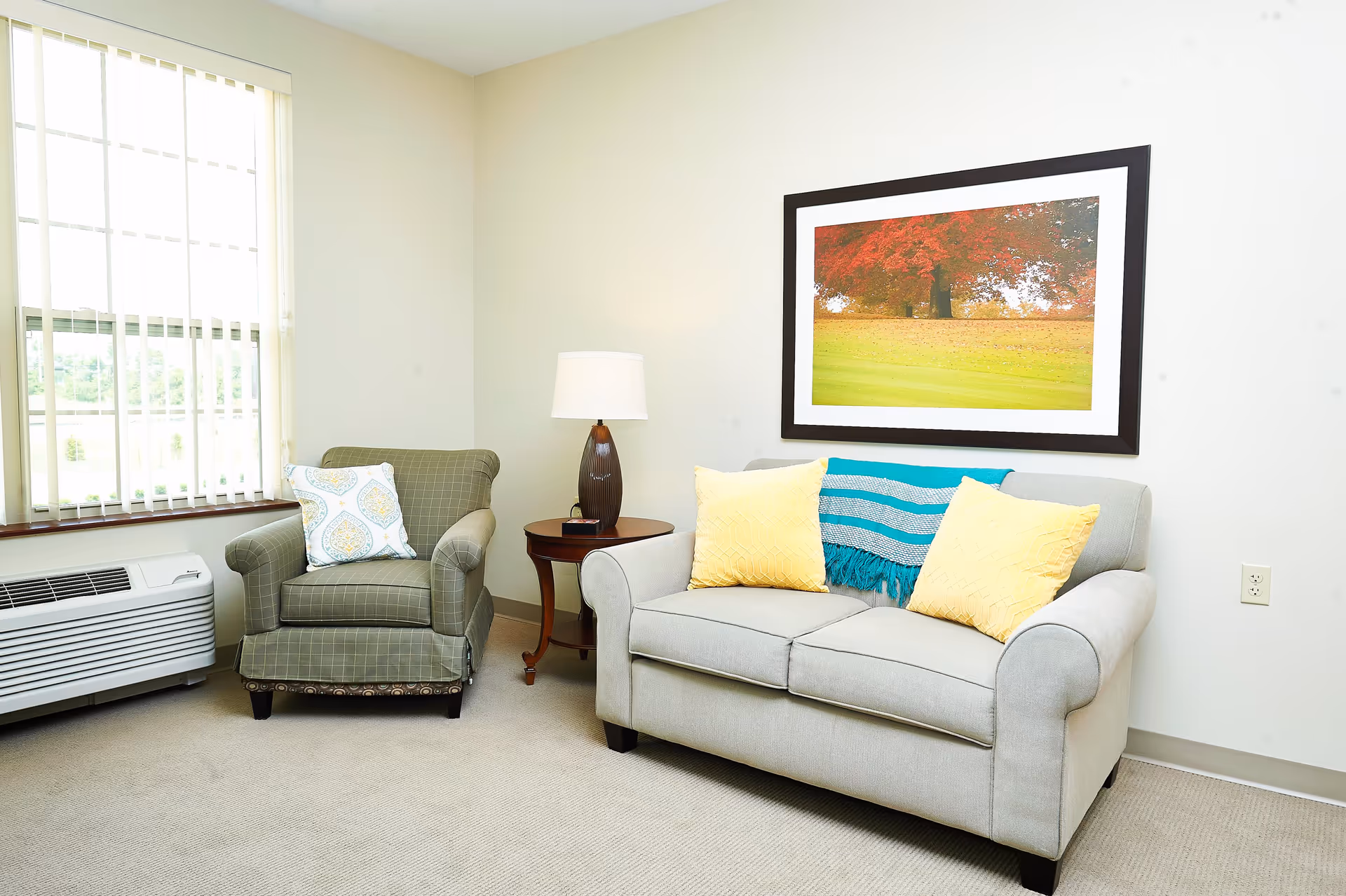 A cozy living room area with a light gray loveseat adorned with two yellow pillows and a blue striped throw blanket. Next to it is a green armchair with a patterned pillow. Between the chairs is a wooden side table with a lamp. A large window with vertical blinds lets in natural light, and a framed picture of a tree with red leaves hangs on the wall.
