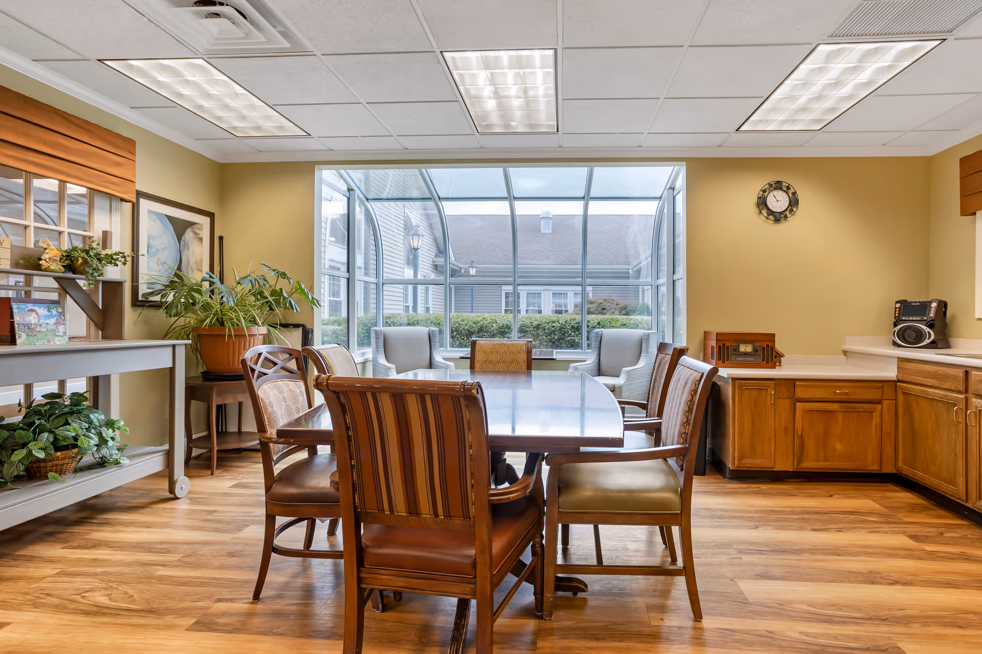 A well-lit room with a wooden dining table surrounded by six chairs of different designs. The room has wood flooring, beige walls, and a large window with a glass enclosure showing an outdoor view of bushes and a building. There are plants on a small table and a shelf, a clock on the wall, and a vintage radio and speaker on the countertop.