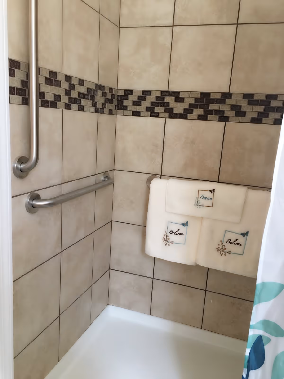 Tiled shower stall with stainless steel grab bars and folded decorative towels hanging on a rail.