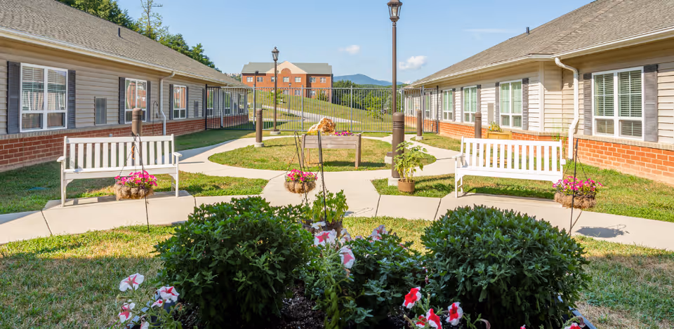Outdoor courtyard area between two single-story buildings with beige siding and brick bases. The courtyard features paved walkways, white benches, hanging flower baskets, potted plants, and a lamppost. In the background, there is a red brick building and a view of distant hills under a blue sky with some clouds.