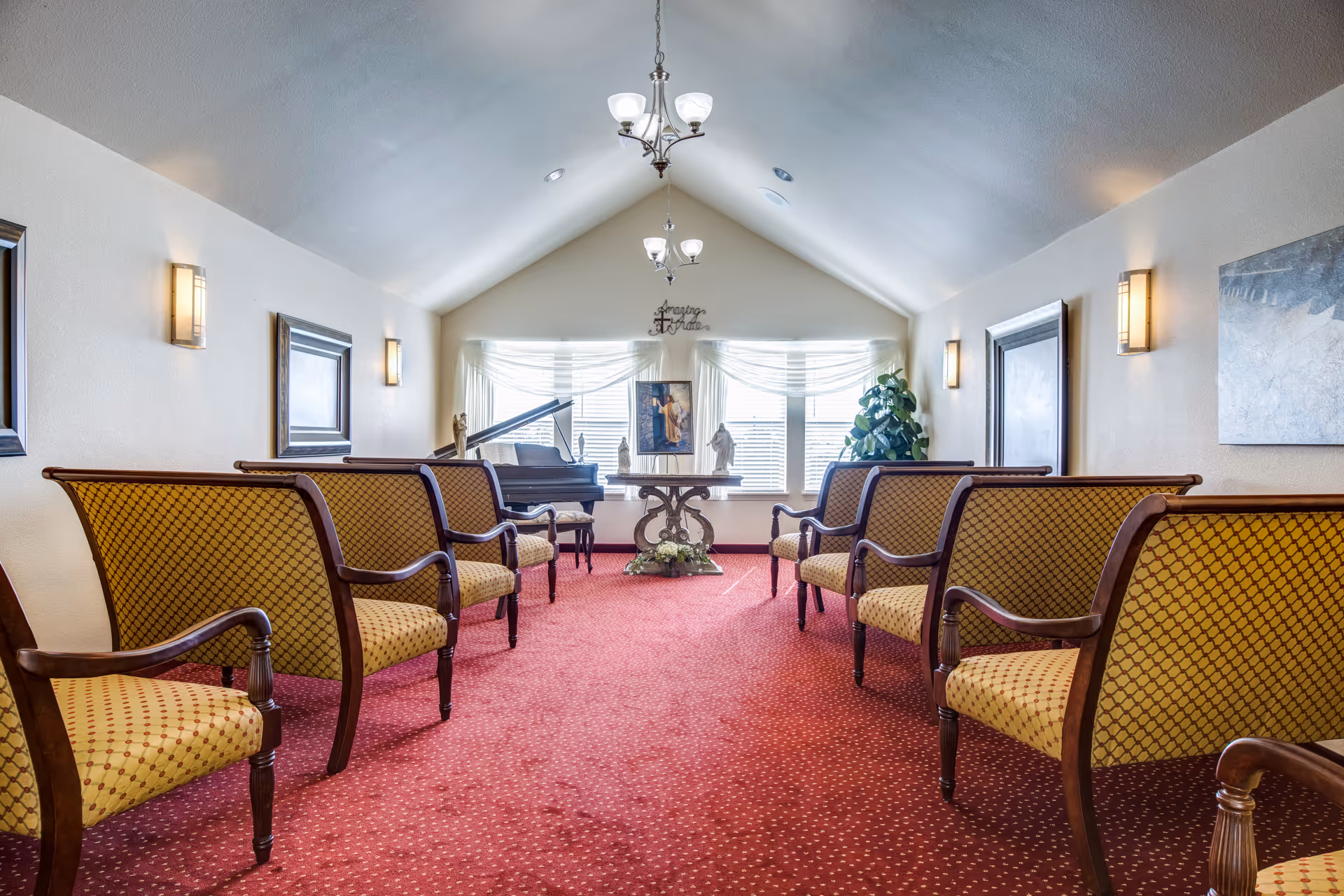Carpeted meeting room with rows of upholstered chairs facing a piano and table under a chandelier.