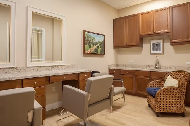 Interior room with beige walls and light wood flooring featuring two large mirrors above a long countertop with wooden cabinets below. There are two gray salon-style chairs in front of the mirrors and a wicker armchair with a blue cushion and a pillow in the corner. Additional wooden cabinets and a sink are mounted on the adjacent wall. A framed picture hangs on the wall between the mirrors and the cabinets.