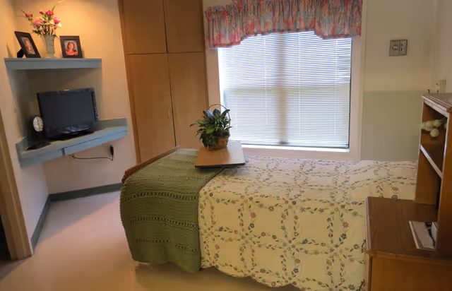A small, cozy bedroom in a senior living facility featuring a single bed with a floral quilt and a green knitted blanket. A wooden tray with a potted plant sits on the bed. The room has a window with closed blinds and floral valance curtains. There is a wall-mounted TV on a corner shelf with framed photos and a clock, and built-in wooden cabinets next to the window.