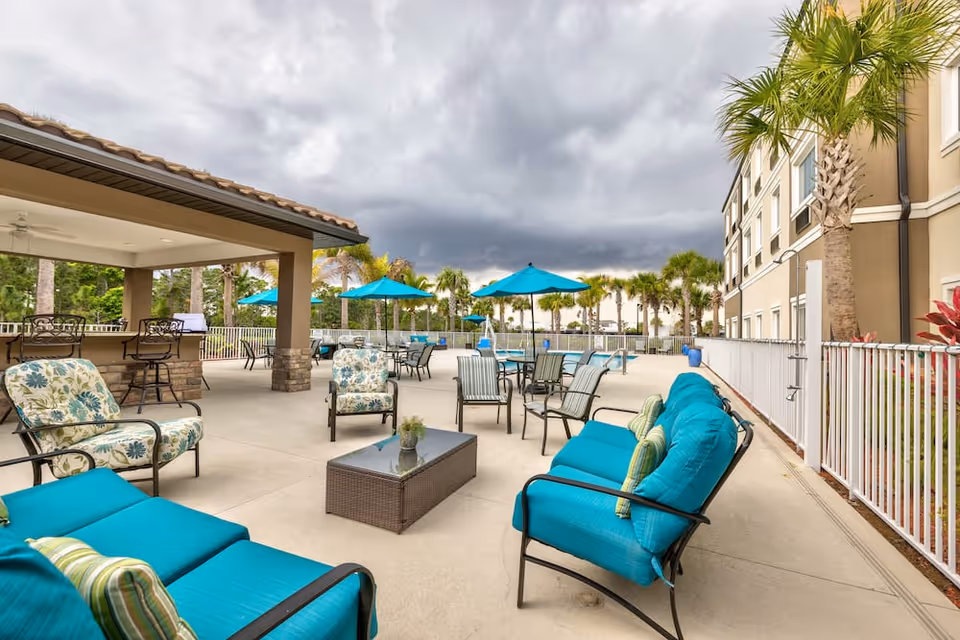 Outdoor patio area with turquoise cushioned chairs, floral patterned chairs, a glass-top coffee table, and several tables with blue umbrellas near a swimming pool. Palm trees and a multi-story building are visible in the background under a cloudy sky.