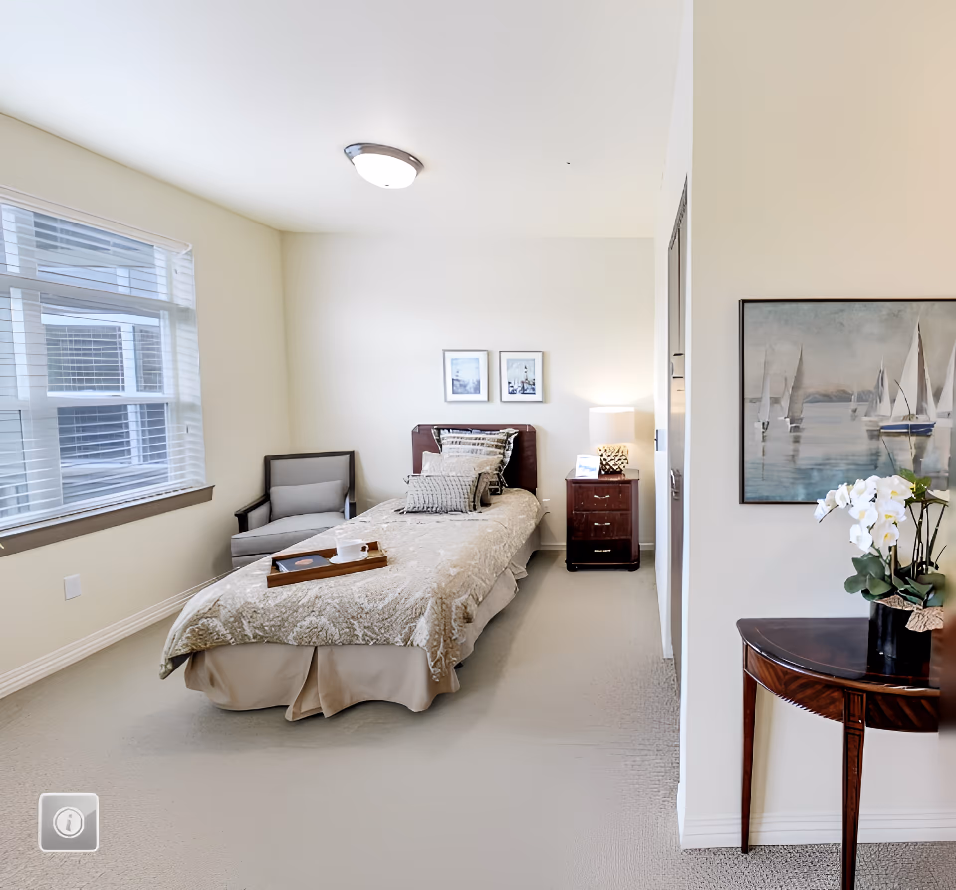 A bright and neatly arranged bedroom in a senior living facility with a single bed covered in a beige patterned bedspread, a wooden nightstand with a lamp and a small framed picture, a gray armchair near a large window with blinds, and a wooden table with a white orchid plant and a painting of sailboats on the wall.