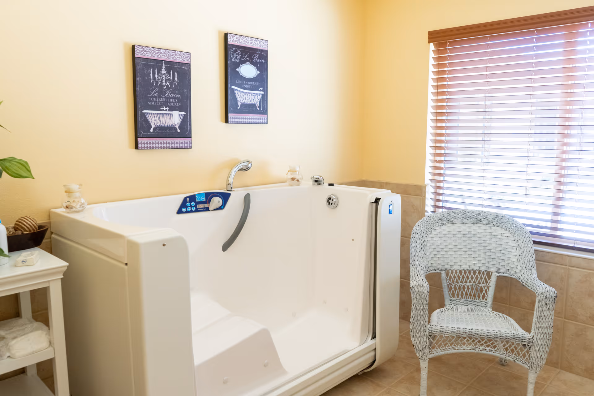 A walk-in bathtub with a built-in seat and faucet in a bathroom with beige walls and tiled floor. Next to the bathtub is a white wicker chair and a small white table with towels and decorative items. Two framed pictures with bathtub illustrations hang on the wall above the bathtub. A window with wooden blinds allows natural light into the room.