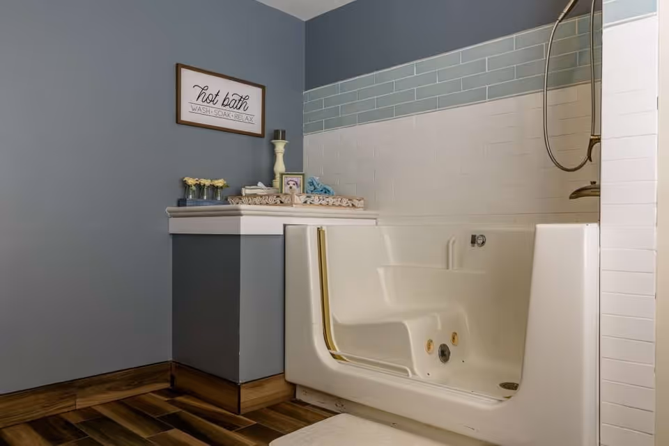 A bathroom with a walk-in bathtub featuring a built-in seat and handrails. The walls are painted gray with white and light blue tile accents. A small counter next to the tub holds decorative items including a candle holder, flowers, and a framed photo. A sign on the wall reads 'hot bath WASH SOAK RELAX.'