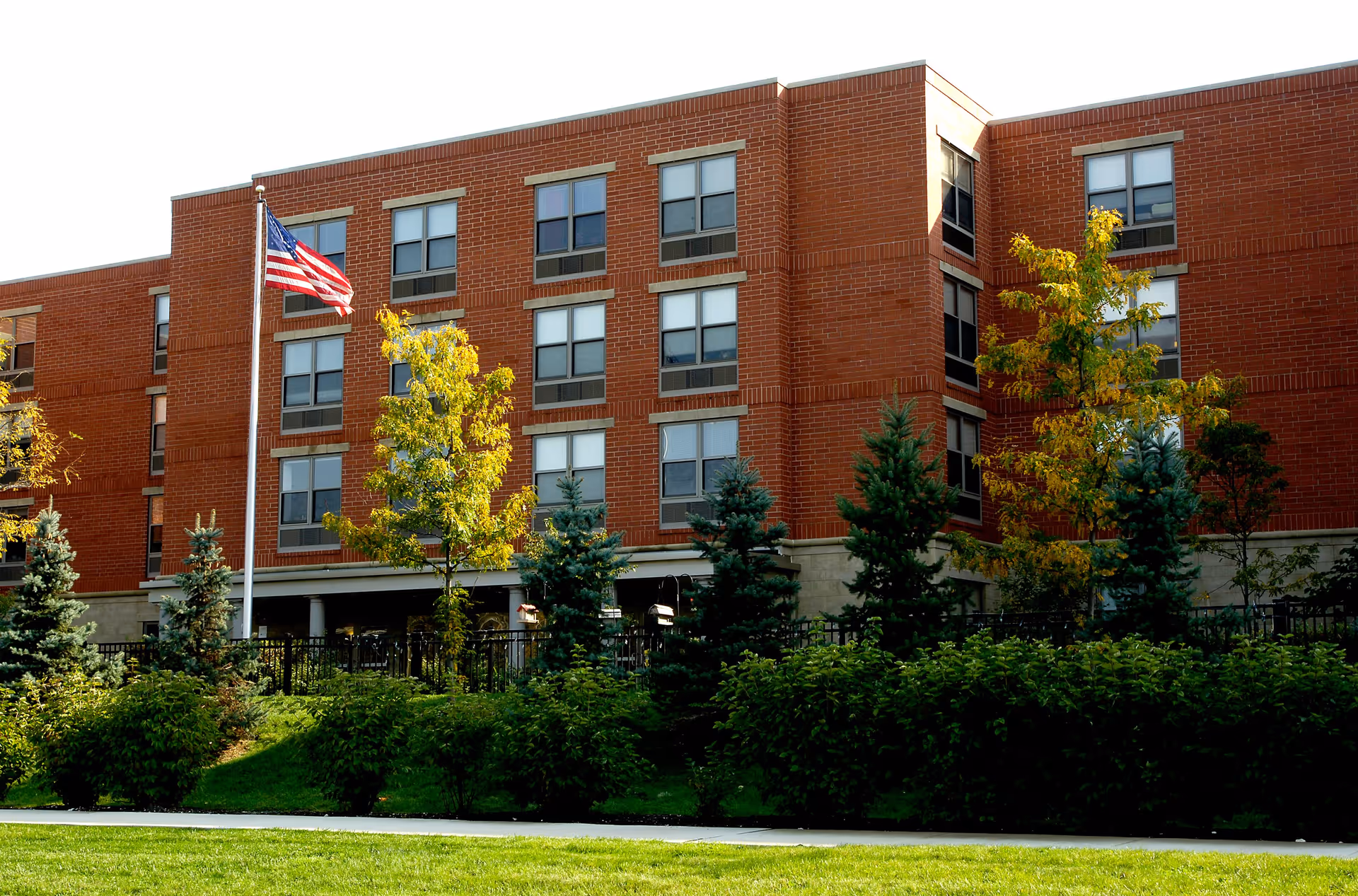 Exterior view of a multi-story brick building with several windows, surrounded by green bushes and trees. An American flag is flying on a flagpole in front of the building under a clear sky.