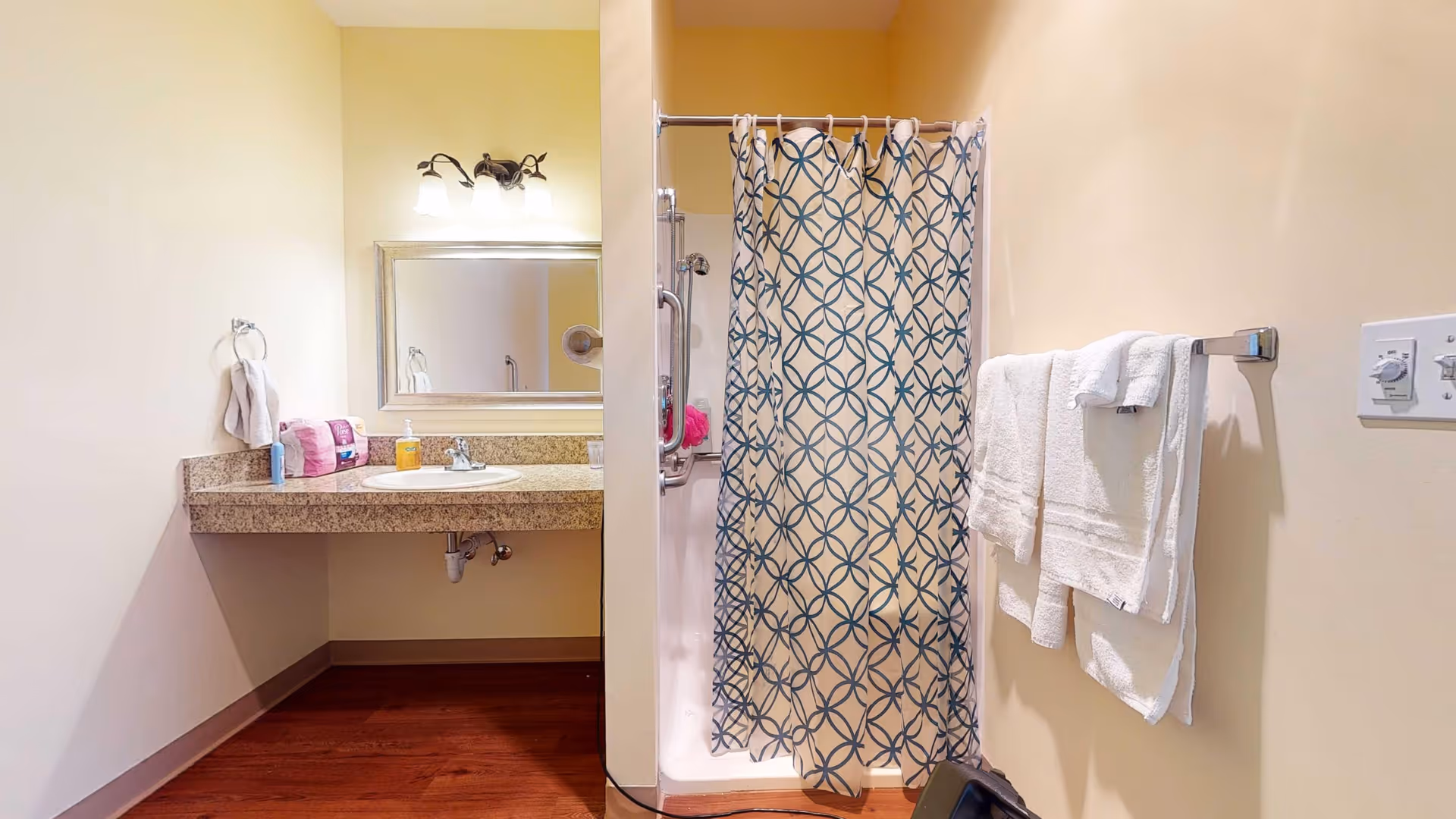 A bathroom with a granite countertop sink on the left, a mirror above the sink, and a light fixture with three bulbs. On the right, there is a shower with a patterned blue and white shower curtain and a towel rack holding white towels. The floor is wooden.