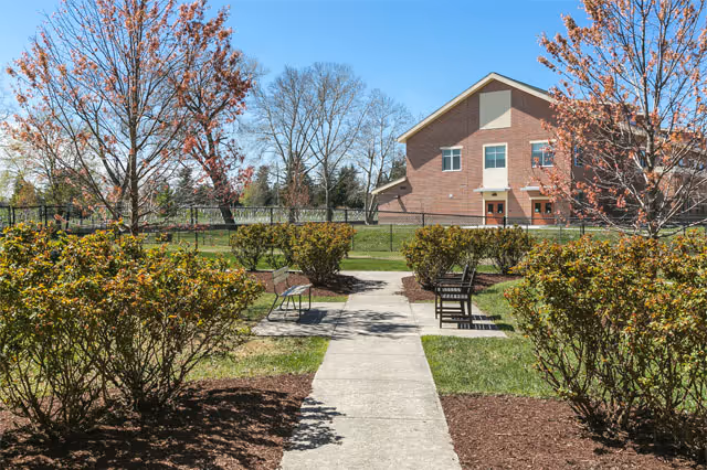 Landscaped outdoor walkway with benches and trimmed bushes leading to a red-brick building under a clear blue sky.